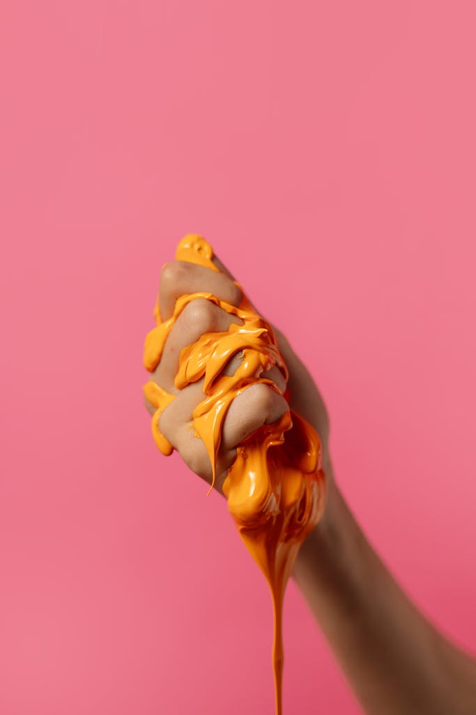 Hand squeezing orange slime against a pink background in a conceptual close-up shot.
